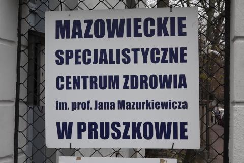 Tablica z napisem "Mazowieckie specjalistyczne centrum zdrowia im. prof. Jana Mazurkiewicza w Pruszkowie" zawieszona na ogrodzeniu