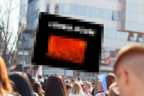 Tłum ludzi na placu w mieście. Ponad ich głowami jest transparent, którego treść jest rozmyta