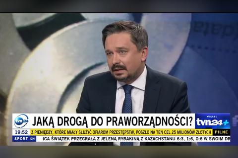 Kadr z programu przedstawiający RPO Marcina Wiącka wypowiadającego się w studiu. Na dole kadru jest pasek z wiadomościami z dużym napisem "Jaką drogą do praworządności?"