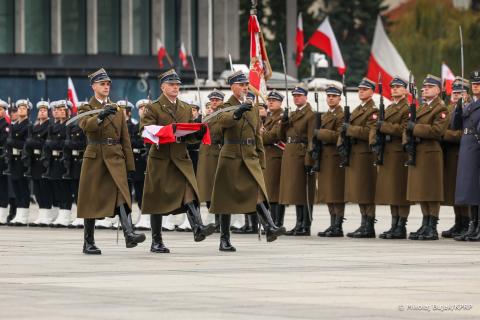 Wzdłuż szeregu żołnierzy stojących na placu maszeruje trzech żołnierzy. Jeden z nich niesie w dłoniach złożoną biało-czerwoną flagę.