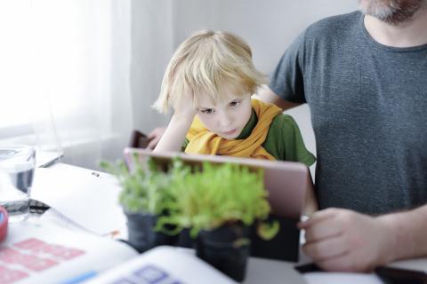 Dziecko siedzi przy stole i trzymając się za głowę wpatruje się w stojący na biurku przed nim tablet. Obok siedzi dorosła osoba.