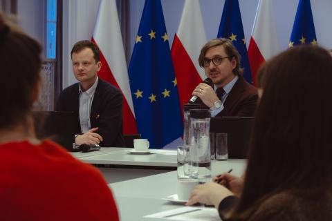 dwie osoby siedzą w sali jedna  przemawia 