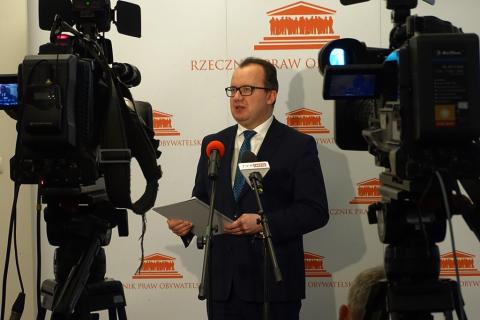 zdjęcie: na pierwszym planie dwie kamery pomiędzy nimi stoi mężczyzna w garniturze