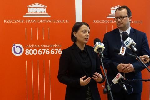 zdjęcie: na pomarańczowym tle widać kobiete w czarnym żakiecie i mężczyznę w garniturze, przed nimi stoją mikrofony