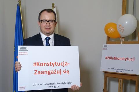 zdjęcie: mężczyzna w garniturze trzyma w ręku tablicę z napisem: Konstytucja - zaangażuj się