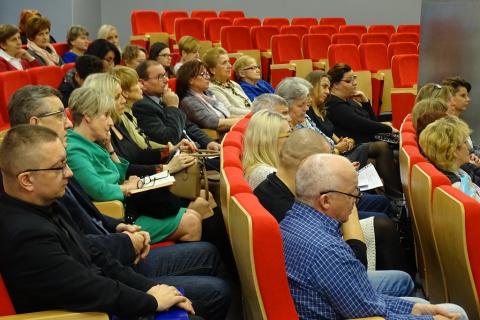 Zdjęcie: ludzie siedzą na czerwonych fotelach w sali audytoryjnej. Ujęcie z boku