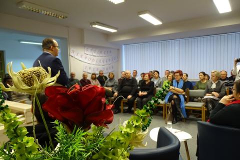 Ludzie w bibliotece siedza na krzesłach ustawionych w rzędach. Mężczyzna stoi i mówi